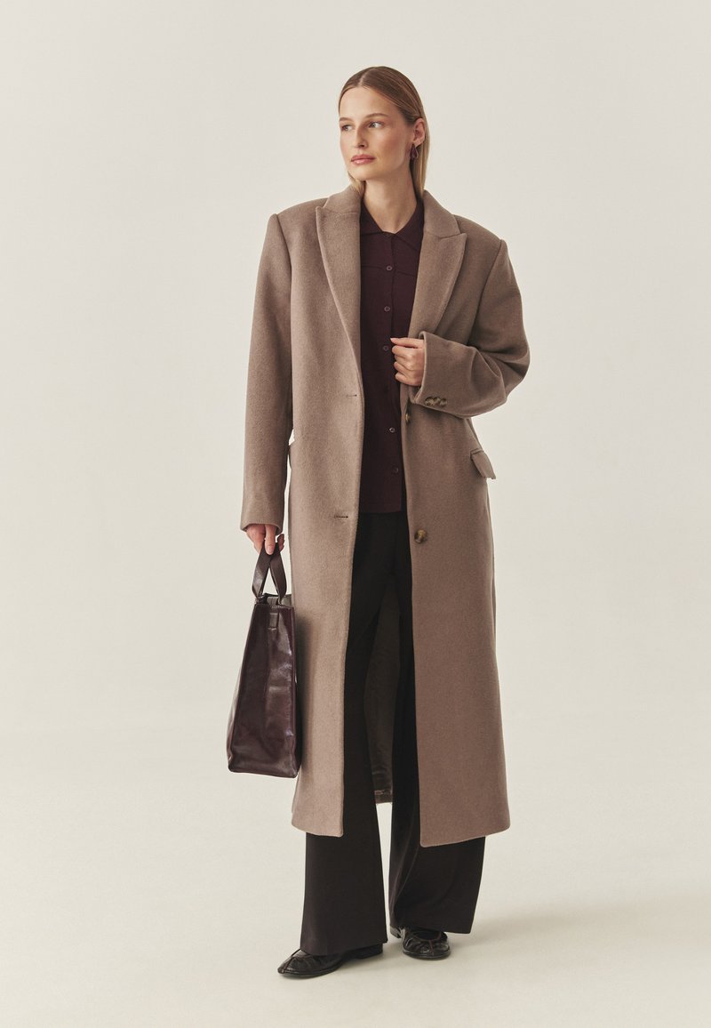 Langer, beiger Wollmantel mit Reverskragen, Ein-Knopf-Verschluss und Seitentaschen. Trägt darunter ein dunkles Hemd und schwarze Hose.