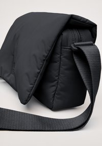 Sacoche noire rembourrée au design élégant, avec un rabat repliable et une bandoulière texturée. Fermeture à zip et forme rectangulaire structurée.