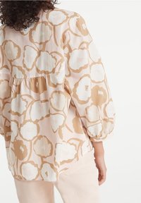 Blusa rosa chiaro con grandi fiori bianchi contornati in beige. Presenta maniche arricciate e un orlo inferiore a balze. Tessuto liscio e leggero.