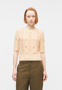 Γυναίκα με κοντά κόκκινα μαλλιά που φοράει ένα ροδακινί πλεκτό πουλόβερ με κοντά μανίκια και πράσινο ελιά παντελόνι ψηλόμεσο, στέκεται μπροστά σε απλό φόντο.