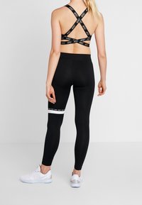 Kvinna vänd bort med svart Nike sportbh med korskorsade band och svarta leggings med vit rand, stående framför vit bakgrund.