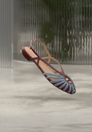 Platte sandaal met bordeauxrode basis, blauwe gevlochten banden in een zonnestraalpatroon, en een dunne enkelband, afgebeeld tegen een vervaagde moderne achtergrond.