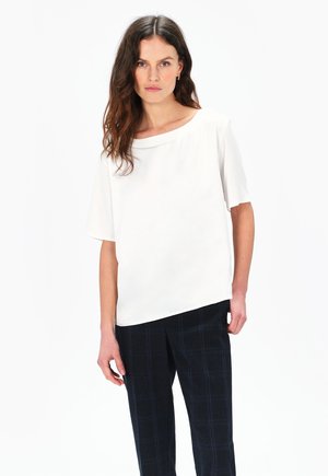 Top bianco a maniche corte con scollatura ampia, abbinato a pantaloni scozzesi scuri. La texture liscia e la vestibilità rilassata creano un aspetto minimalista.