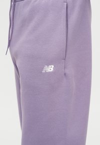 Κοντινό πλάνο μοβ φόρμας με πλαϊνή τσέπη και λευκό λογότυπο New Balance στο μηρό.