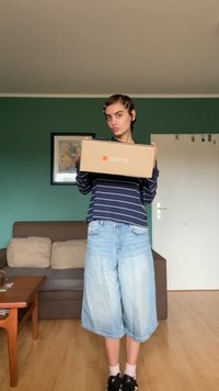 Papist kast, millel on Zalando logo, hoitud kätes. Isik kannab tumesinist triibulist pika varrukaga särki ja helesiniseid laiad lühikesi pükse ning tumedat jalatsit.
