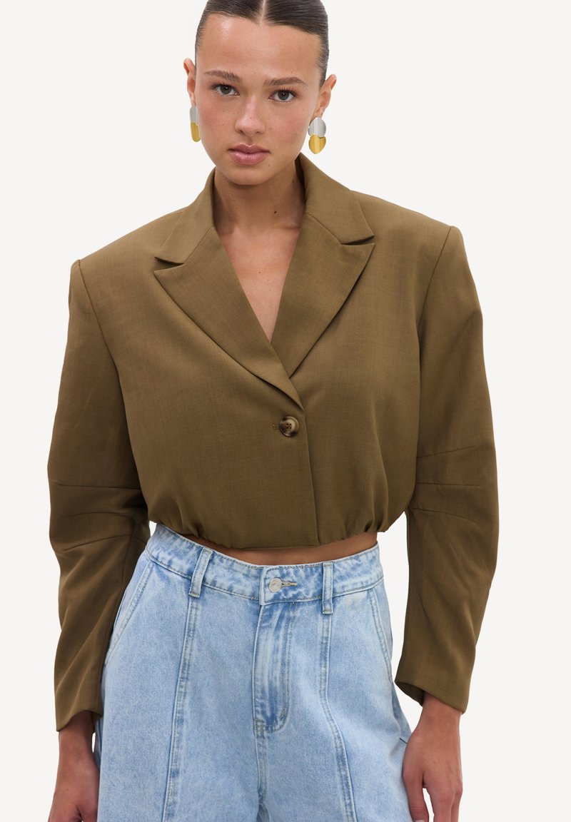 Femme portant un blazer court olive à un bouton et un jean taille haute bleu clair, associé à des boucles d'oreilles pendantes bicolores.