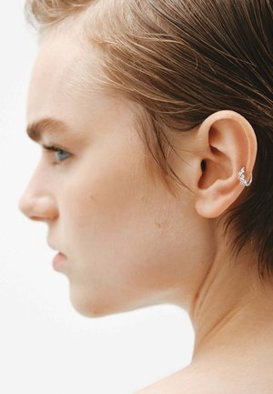 Pendiente de aro plateado con pequeños acentos geométricos, que presenta un acabado liso. El modelo tiene el cabello corto, ligeramente ondulado y un tono de piel neutro.