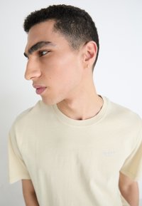 Beige katoenen t-shirt met een ronde hals, korte mouwen en een klein lichtblauw logo op de borst. Model toont een zijprofiel.