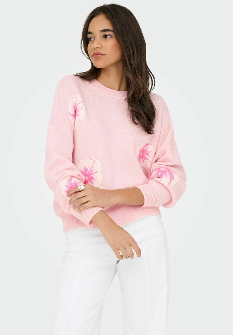 Pull en tricot rose clair avec accents floraux en rose plus foncé. Le design inclut un col rond et une coupe ample, légèrement surdimensionnée.