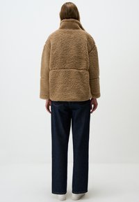 Bruine nepbontjas met een hoge kraag, oversized pasvorm en wijde mouwen, gecombineerd met een donkere spijkerbroek en witte sneakers.