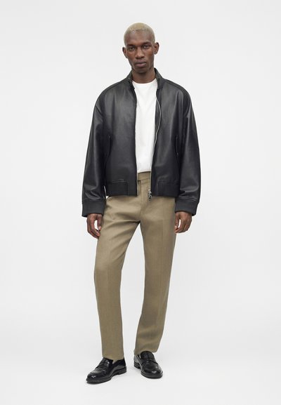 Άνδρας στέκεται φορώντας μαύρο δερμάτινο μπουφάν πάνω από άσπρο πουκάμισο, μπεζ παντελόνι και μαύρα δερμάτινα παπούτσια σε λευκό φόντο.