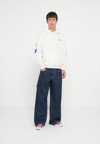 Hoodie branco com bolso frontal e logótipo colorido, combinado com calças amplas de riscas azuis e sapatilhas com detalhes em vermelho.