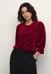 Blusa de terciopelo rojo con rayas verticales, cuello redondo y mangas abullonadas hasta el codo, combinada con pantalones negros. Textura suave y ajuste relajado.
