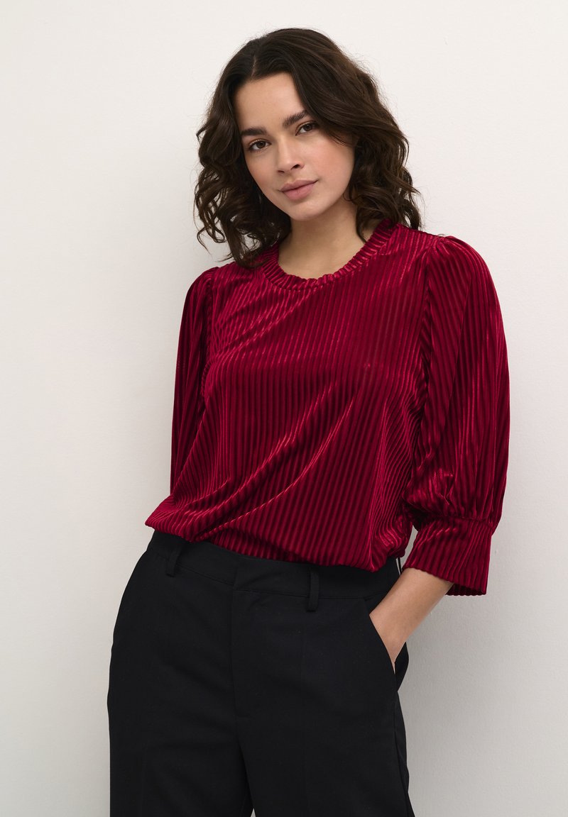 Blusa de terciopelo rojo con rayas verticales, cuello redondo y mangas abullonadas hasta el codo, combinada con pantalones negros. Textura suave y ajuste relajado.