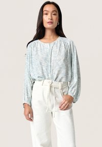 Blusa azzurro chiaro con un motivo ondulato, bottoni davanti e maniche arricciate. Abbinata a pantaloni bianchi a vita alta con texture.