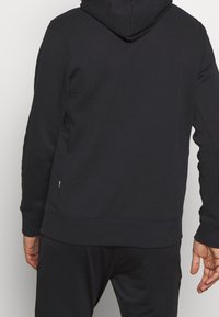 Sweat à capuche noir, tissu doux, avec capuche et poignets côtelés. Design sans coutures avec des surpiqûres discrètes et une petite étiquette logo à l'ourlet.