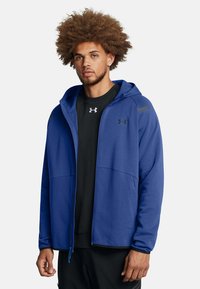 Blaue Zip-Hoodie aus einem weichen, glatten Stoff. Verfügt über eine Kapuze, Fronttaschen und schwarze Akzente an den Schultern und im Logo.