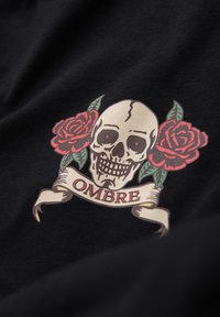 Crâne aux yeux rouges centré entre deux roses rouges avec des feuilles vertes au-dessus d'un parchemin portant l'inscription « OMBRE » sur un tissu noir.
