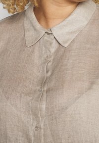 Světle hnědá lněná košile s límečkem a zapínáním na knoflíky. Textura je mírně průhledná, s jemnými variacemi v látce.