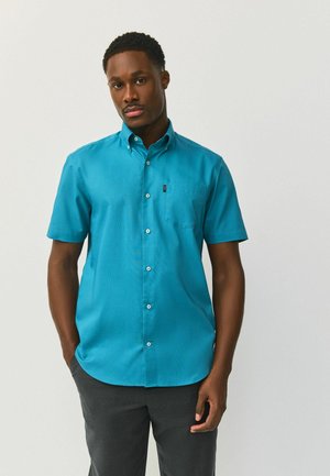 Muž v tyrkysovej krátkorukávovej košeli s gombíkmi a tmavosivých nohaviciach, stojaci s rukami vreckách na jednoduchom svetlom pozadí.