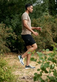 Tan short-sleeve shirt, black athletic shorts, and grey-green athletic shoes with yellow accents, walking on uneven ground surrounded by greenery.
