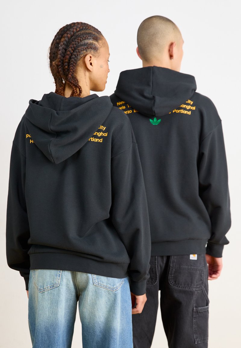 Czarne bluzy z kapturem o luźnym kroju, z żółtym napisem i zielonym logiem na plecach. Miękki materiał o gładkiej teksturze, zestawione z dżinsowymi spodniami.