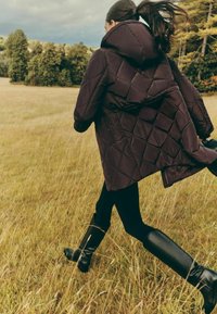 Tikitud tumelilla vurrutatud jope kapuutsiga, millel mustad säärised ja põlveni ulatuvad mustad saapad, taustal rohulõikamise väli.