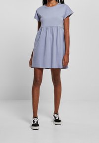 Frau trägt ein helllila kurzärmeliges Babydoll-Kleid mit gerafftem Bund und schwarzen Low-Top-Sneakern mit weißen Schnürsenkeln.