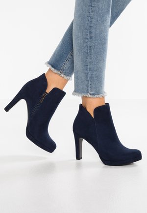 Pieds chaussés de bottines à talons hauts en daim bleu foncé avec des fermetures éclair latérales, associées à un jean skinny bleu clair à ourlet effiloché.
