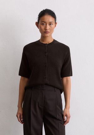Dunkelbraune kurzärmelige Strickjacke mit gerippter Textur, Rundhalsausschnitt und Knopfverschluss. Kombiniert mit schwarzen, hochgeschnittenen Hosen.