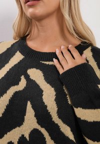 Blond kvinna som bär en svart och beige mönstrad stickad tröja, med handen vilande på bröstet, som visar långa naglar och läppar.