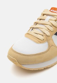 Beige and white athletic sneaker featuring a suede and mesh upper, round toe, flat laces, and textured rubber sole for grip.