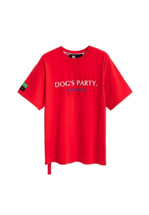 Mujer rubia vestida con una camiseta roja «DOG'S PARTY» de gran tamaño, pantalones cortos blancos, calcetines blancos, zapatos beige y sosteniendo una etiqueta larga de tela.