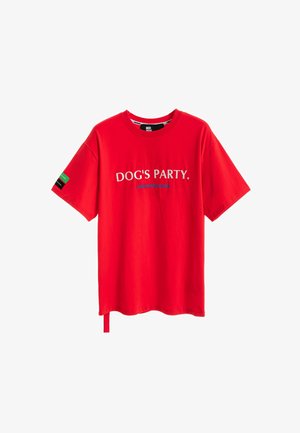 Mujer rubia vestida con una camiseta roja «DOG'S PARTY» de gran tamaño, pantalones cortos blancos, calcetines blancos, zapatos beige y sosteniendo una etiqueta larga de tela.