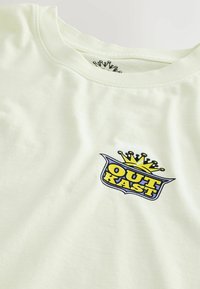 Vaaleankeltainen puuvillapaita, jossa on sininen ja keltainen graafinen logo, ja tekstin "OUT KAST" ylle on sijoitettu kruunusuunnittelu. Pyöreä kaula-aukko.