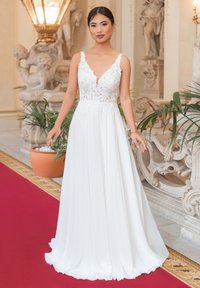 Abito da sposa bianco con bustier in pizzo e scollatura a V profonda, gonna fluente e dettagli floreali, il tutto su uno sfondo ornato.
