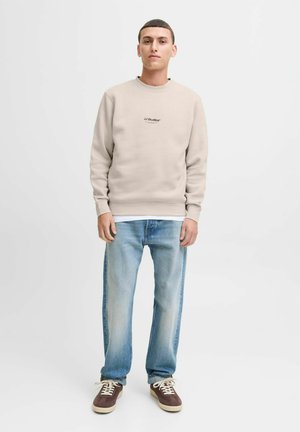Man som bär en beige JJ Studios sweatshirt, ljusblå jeans och bruna sneakers står mot en enfärgad vit bakgrund.