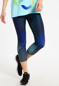 Leggings Capri avec un motif géométrique bleu et vert, fabriqués à partir d'un matériau extensible avec des rayures contrastées, associés à des baskets noires.