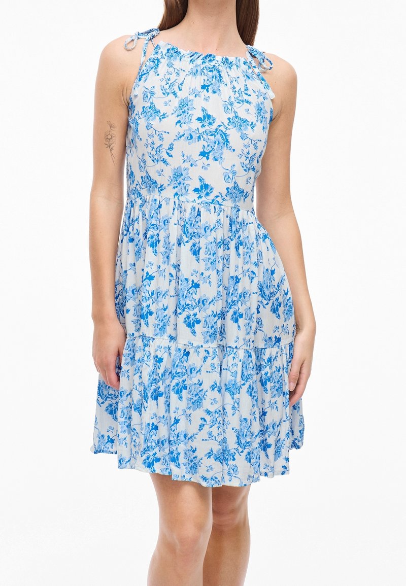Femme portant une robe blanche sans manches à imprimé floral bleu avec des bretelles à nouer, debout avec les bras relâchés le long du corps.