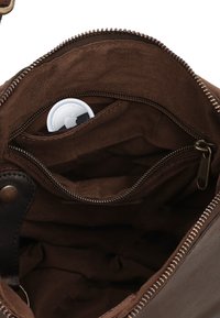 Hnedá kožená taška otvorená, aby ukázala tmavý textilný interiér s malým vreckom na zips obsahujúcim biely Apple AirTag.