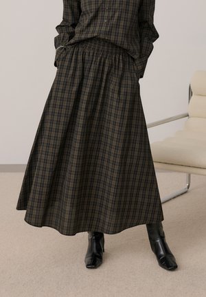 Femme portant une chemise à carreaux foncée boutonnée et une jupe longue assortie avec des bottes en cuir noires à talons, debout à l'intérieur près d'une chaise crème.