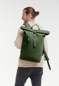 Mochila verde tipo roll-top hecha de material duradero, con un bolsillo frontal con cremallera, correas ajustables y una impresión de logo.