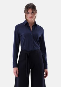 Navy long-sleeve shirt with button closure and pointed collar. Soft, smooth fabric with a fitted design, paired with wide black trousers and belt.