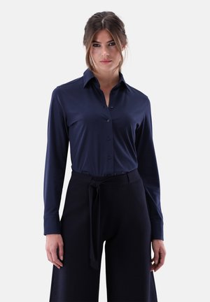 Camicia a maniche lunghe color blu navy con chiusura a bottoni e colletto appuntito. Tessuto morbido e liscio con design aderente, abbinata a pantaloni neri larghi e cintura.