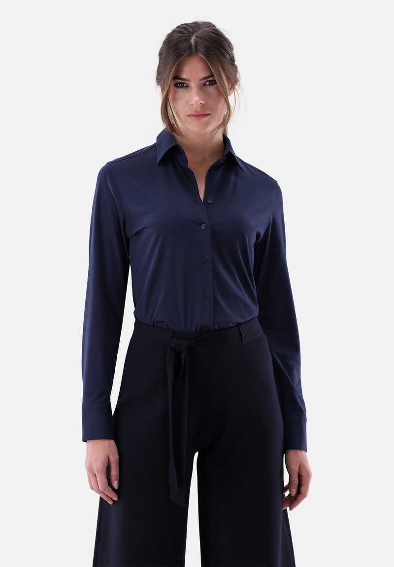 Navy long-sleeve shirt with button closure and pointed collar. Soft, smooth fabric with a fitted design, paired with wide black trousers and belt.