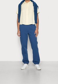 Chemise jaune clair à manches courtes, pantalon de train bleu avec des manches élastiques, baskets blanches, et un pull bleu foncé drapé sur les épaules.