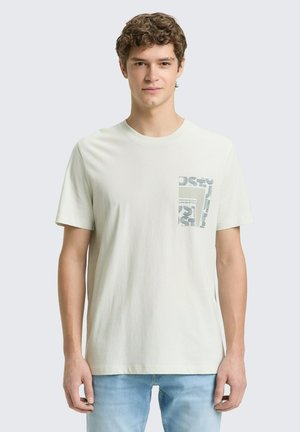 Junger Mann trägt ein schlichtes weißes T-Shirt mit einem gemusterten quadratischen Design auf der linken Brust, kombiniert mit hellblauen Jeans.