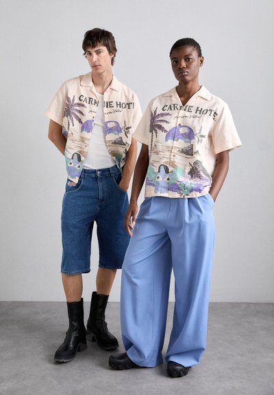Two models wear casual outfits. One has a patterned short-sleeve shirt, denim shorts, and black boots. The other wears a light blue loose-fit pant.