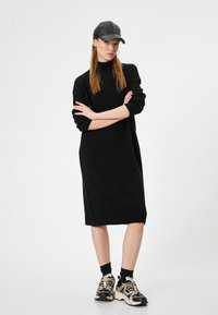 Abito nero lavorato a maglia con collo alto e orlo a coste. La modella indossa un berretto nero e sneakers eleganti con dettagli dorati. Outfit casual.