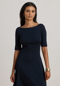 Abito blu navy con scollatura a barca e maniche a tre quarti, caratterizzato da una texture liscia e spacco laterale per una maggiore libertà di movimento. Bracciali dorati accentuano il polso.